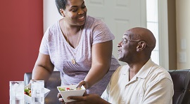 A mid adult African-American woman in her 30s at home with her father, a senior man in his 60s. He is sitting at the dining room table and she is serving bowls of salad.
