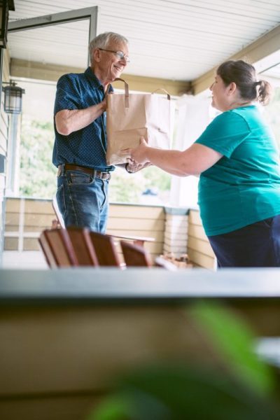 A kind and caring neighbor or friend delivers fresh produce from the grocery store to an elderly man at his home.  He receives the gift with a smile on his face, grateful for the help and assistance.  Vertical image with copy space.